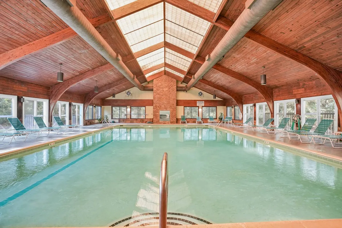 An inviting indoor swimming pool illuminated by natural light streaming through large windows, with wooden beams adding warmth and character. The calm water and spacious pool deck provide a tranquil setting for swimming and socializing, reflecting the resort’s focus on year-round wellness and relaxation.