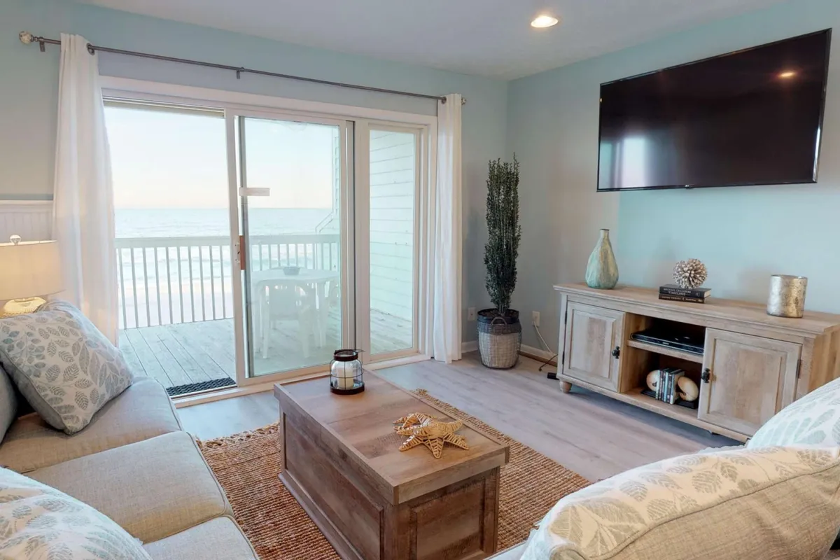 A tranquil living room with a large sliding glass door leading to the balcony, a comfortable sofa with patterned cushions, a decorative coffee table adorned with a starfish, and a wooden TV unit with coastal decor. Through the open doors, the ocean views are visible, connecting the interior seamlessly with the outdoors.