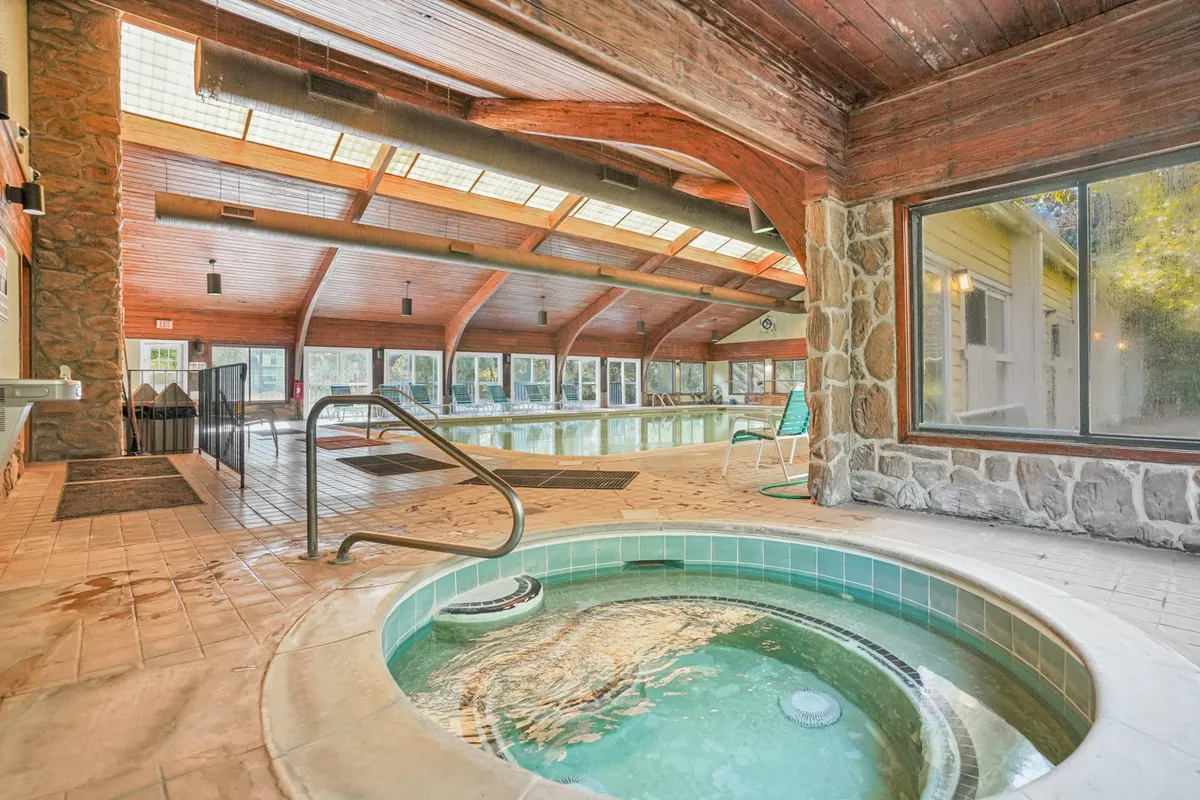 A serene interior view featuring a luxurious hot tub adjacent to an indoor pool, framed by expansive windows and rustic wooden beams. The setting exudes relaxation with ample space for guests to unwind, socialize, or enjoy peaceful moments by the water.
