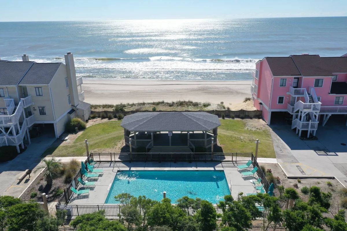 The scene highlights a sparkling swimming pool set beside the beach, with poolside seating, surrounding resort buildings, and a gazebo. The close proximity to the shoreline and inviting pool create a resort-style, all-ages-friendly vibe.