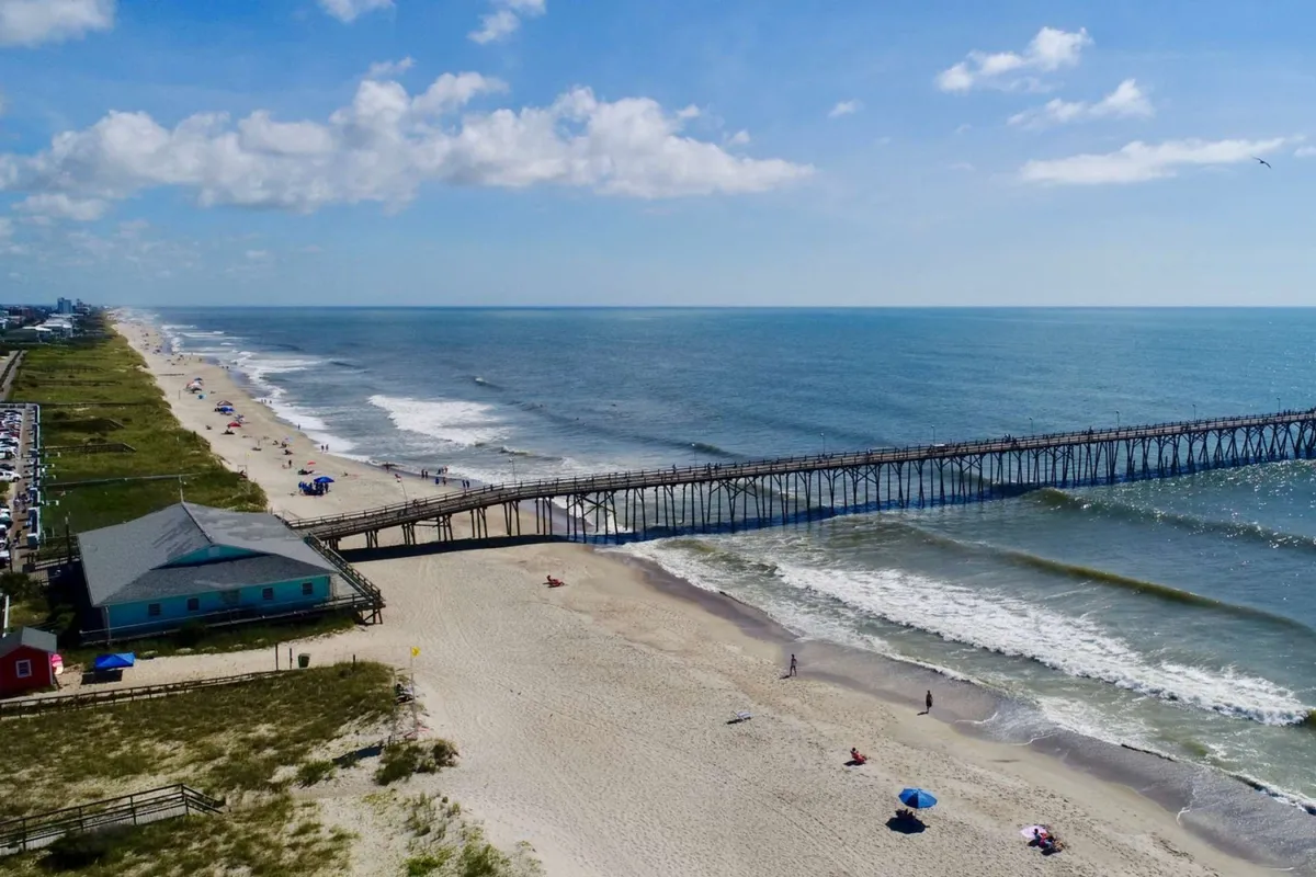 This exterior image captures a bustling beach scene with families and children playing in the sand, colorful umbrellas dotted along the shore, and a pier stretching into the ocean. The tranquil sky and gentle ocean waves create an inviting atmosphere for beach activities.