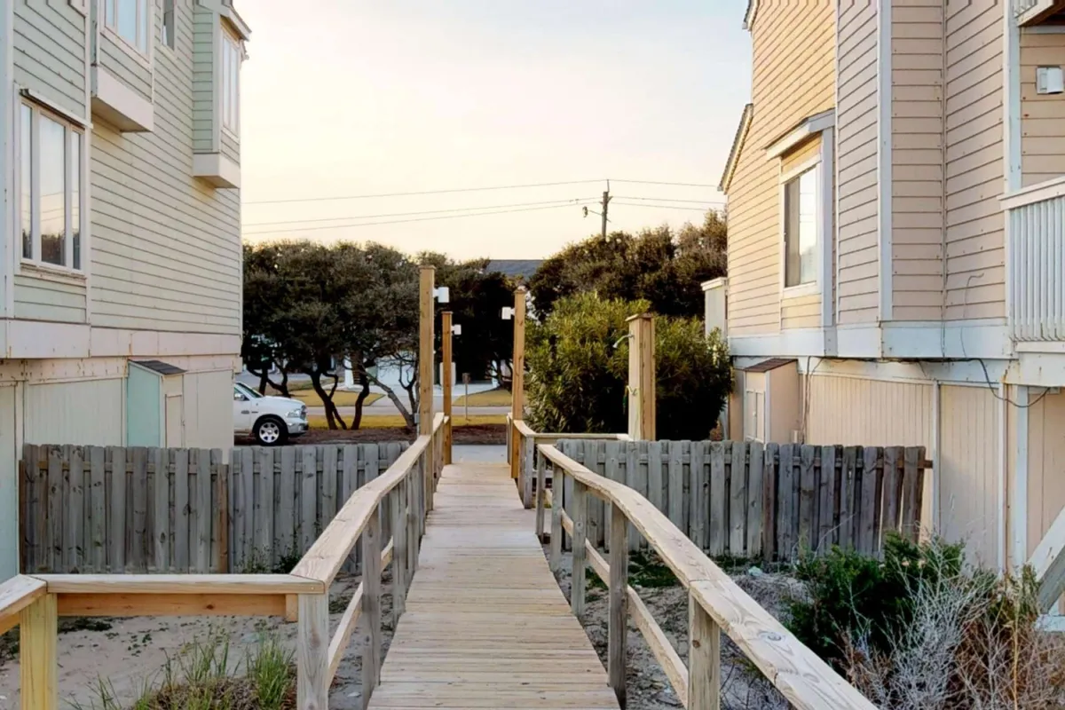 This image shows a serene wooden walkway flanked by lush greenery, leading toward the facades of resort buildings. The inviting path hints at easy beach access and opportunities for peaceful walks, with the resort community visible in the background.