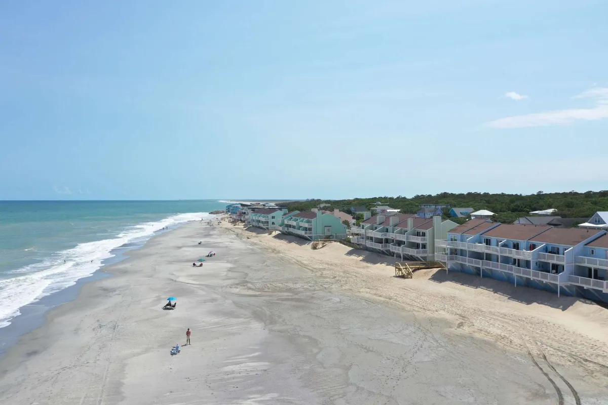 A broad, inviting beach with gentle ocean waves and people relaxing on the sand in front of beachfront properties. The scene suggests active engagement with the shoreline, from sunbathing to casual walks along the water, underlining the vacation vibe.