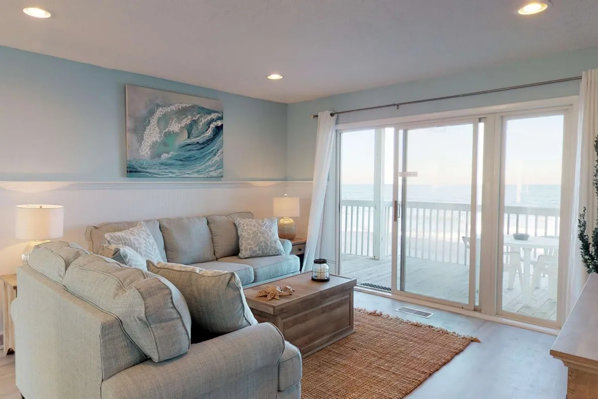An inviting living space with a plush sofa, a coffee table, and an unobstructed ocean view through wide sliding doors. Artwork featuring a wave and tasteful decor complement the airy, serene atmosphere.