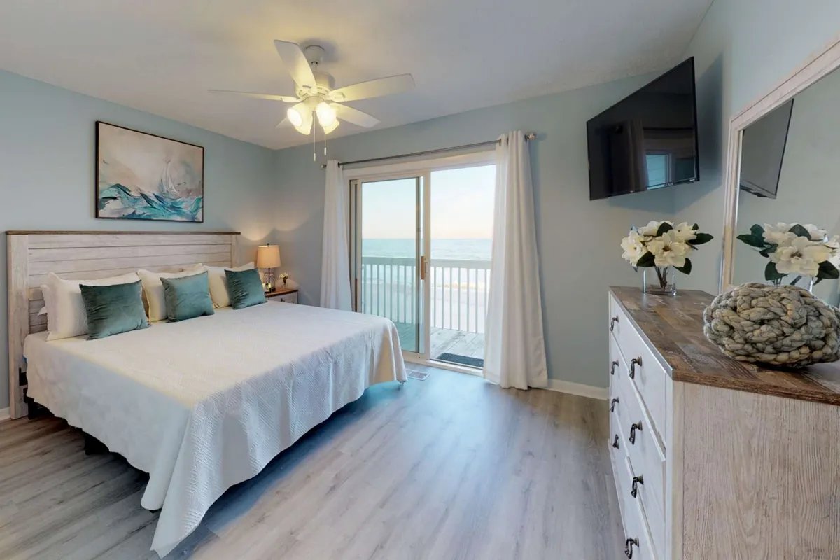 A bright and inviting bedroom with a large bed, dressed in crisp white linens and soft accent pillows. A sliding glass door opens to a balcony, letting in natural light and gentle ocean breezes. Decorative artwork and a dresser add thoughtful touches, emphasizing comfort and tranquility.
