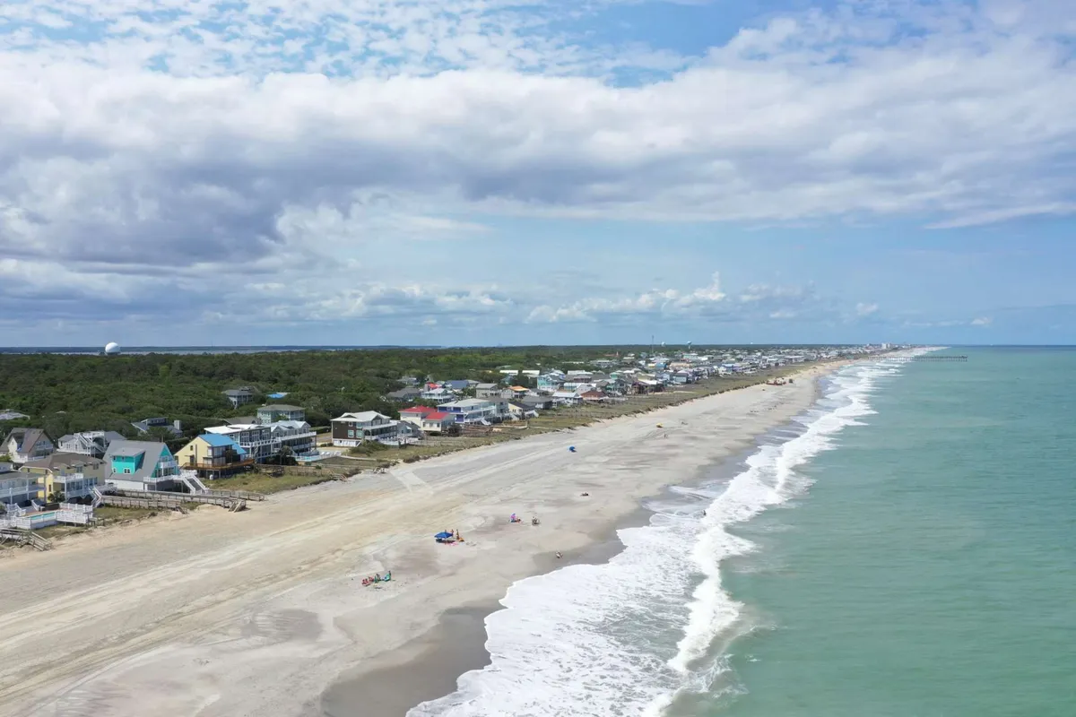 A coastal view capturing wide stretches of sandy beach, gentle waves, nearby houses, and blue sky overhead. It suggests opportunities for surfing, biking, and water sports, with easy access to local shops and eateries visible in the background.