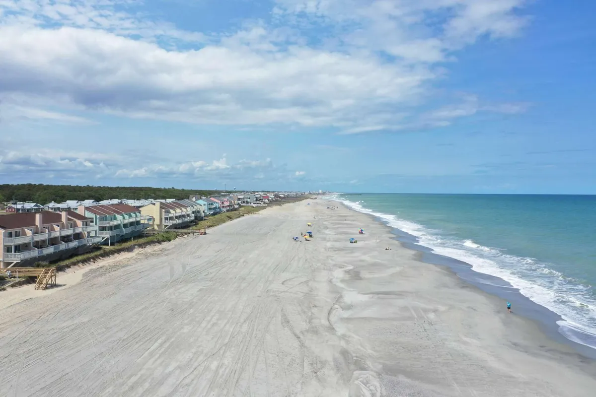 A vibrant exterior scene featuring beachfront houses, an expansive sandy beach, rolling ocean waves, and people enjoying the shoreline. The activity and scale evoke a lively coastal vibe, perfect for swimming, beach walks, and sunbathing.